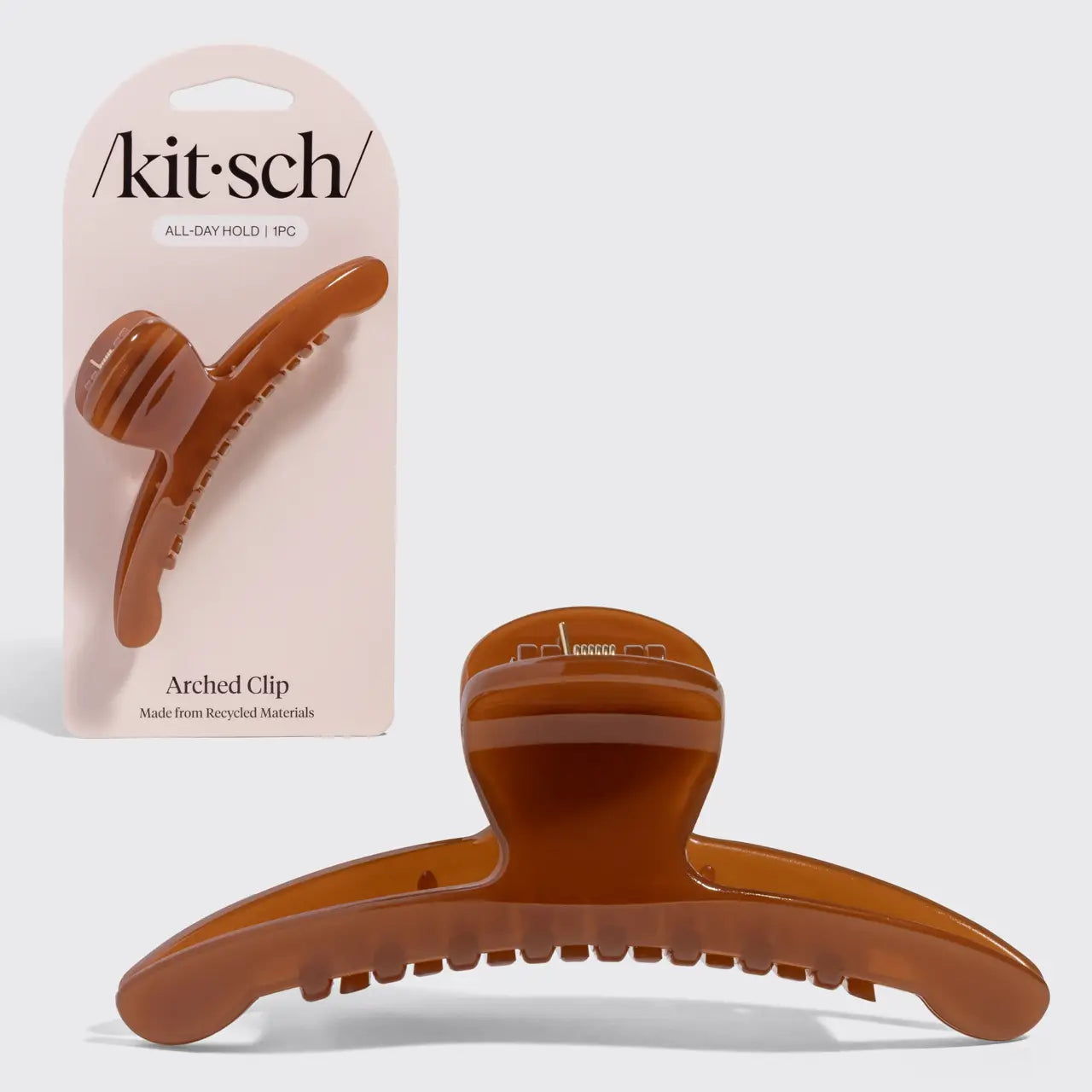 A translucent amber-brown arched hair claw clip shown in front of its light pink Kit-sch retail card packaging on a light gray background.
