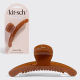 A translucent amber-brown arched hair claw clip shown in front of its light pink Kit-sch retail card packaging on a light gray background.