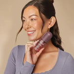 A smiling woman with dark hair in a bun holds a Kitsch Soft Santal Hair Perfume against her neck, wearing a lavender long-sleeve top against a solid beige background.