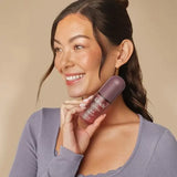A smiling woman with dark hair in a bun holds a Kitsch Soft Santal Hair Perfume against her neck, wearing a lavender long-sleeve top against a solid beige background.