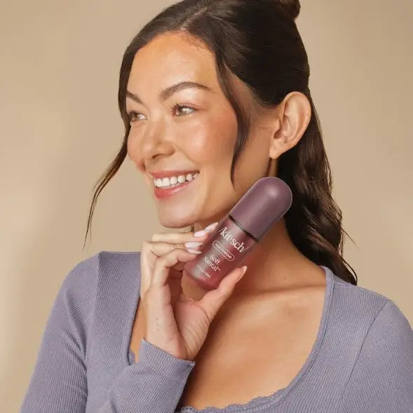 A smiling woman with dark hair in a bun holds a Kitsch Soft Santal Hair Perfume against her neck, wearing a lavender long-sleeve top against a solid beige background.