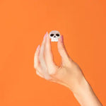 A hand with pink manicured nails holds a small white skull-shaped accessory against a solid orange background.