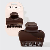 Two dark brown glossy claw hair clips shown on a light gray background, with one clipped to a pale pink Kit·sch card and one loose in front. The Kit·sch brand name is visible on both the card and the clips. 