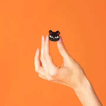A hand holding a small black cat-shaped ring against a solid orange background.