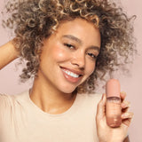 Woman holding a bottle of Kitsch makeup product against a pink background