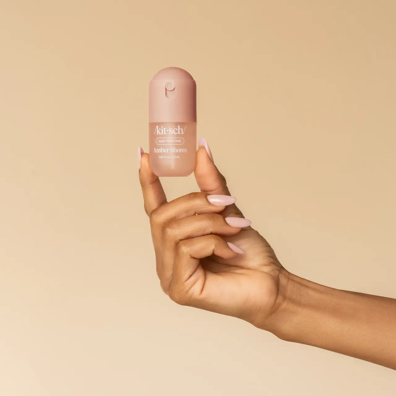 A hand with pale pink manicured nails holds a small bottle of Kitsch Mini Amber Shores hair Perfume against a solid beige background.