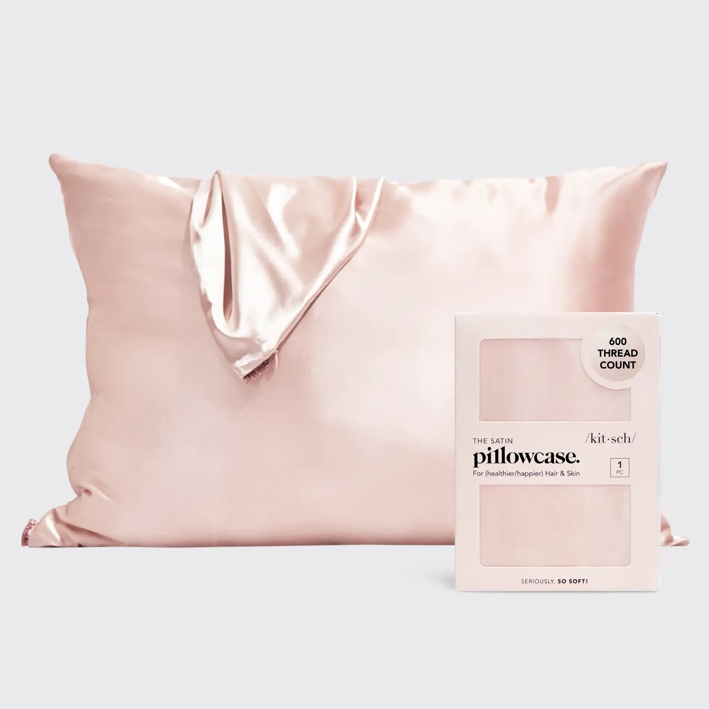 A pale blush pink satin pillowcase with a draped fold, shown next to its product packaging box on a light gray background. The box is labeled 'the silk pillowcase' and 'kit-schi'.