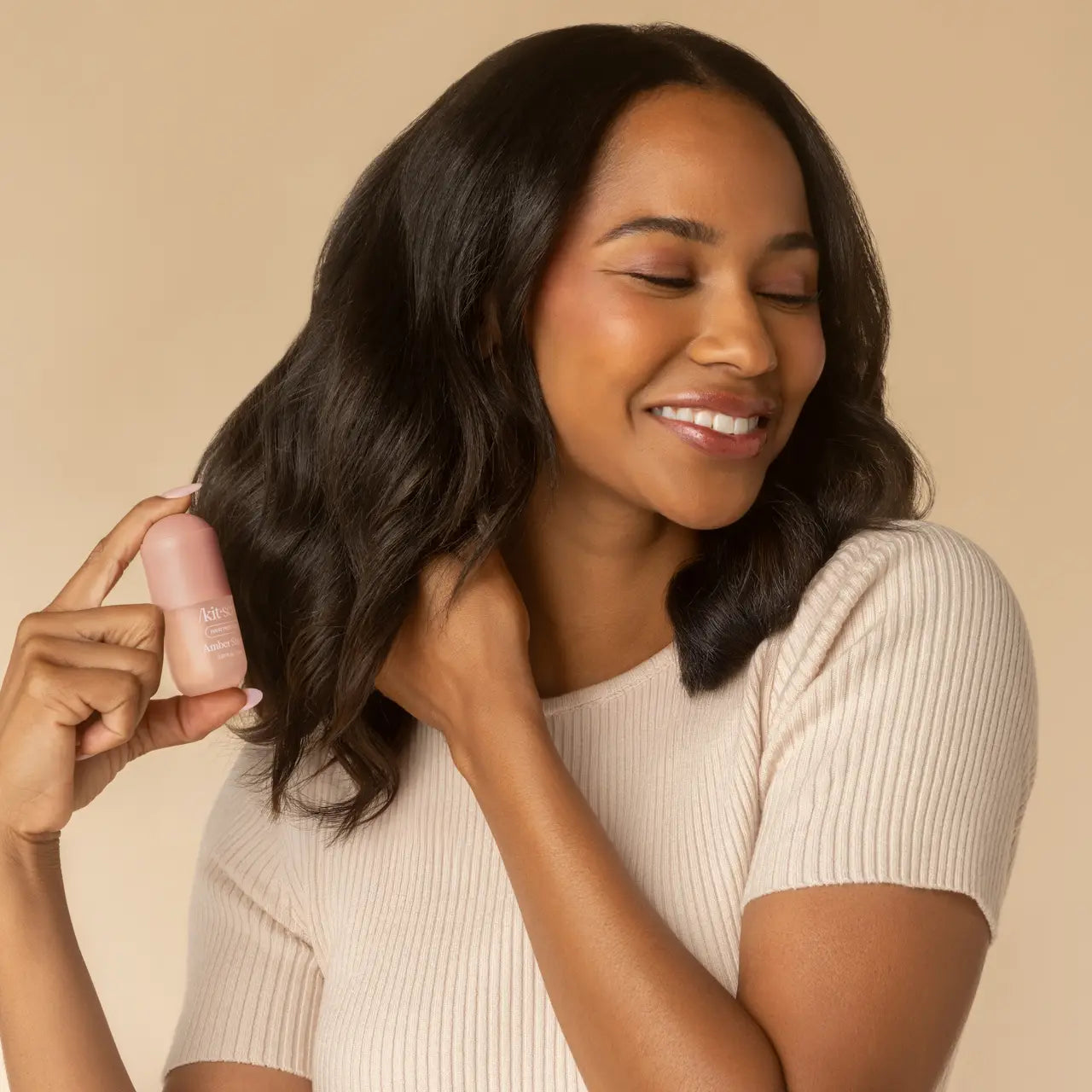 A woman with long, wavy dark brown hair holds a small Kitsch Mini Amber Shores Hair Perfume against a warm beige studio background.