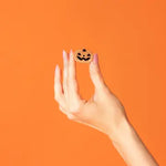 A hand with pink manicured nails holds a small round wooden pumpkin cookie with a black jack-o'-lantern face against a solid orange background.