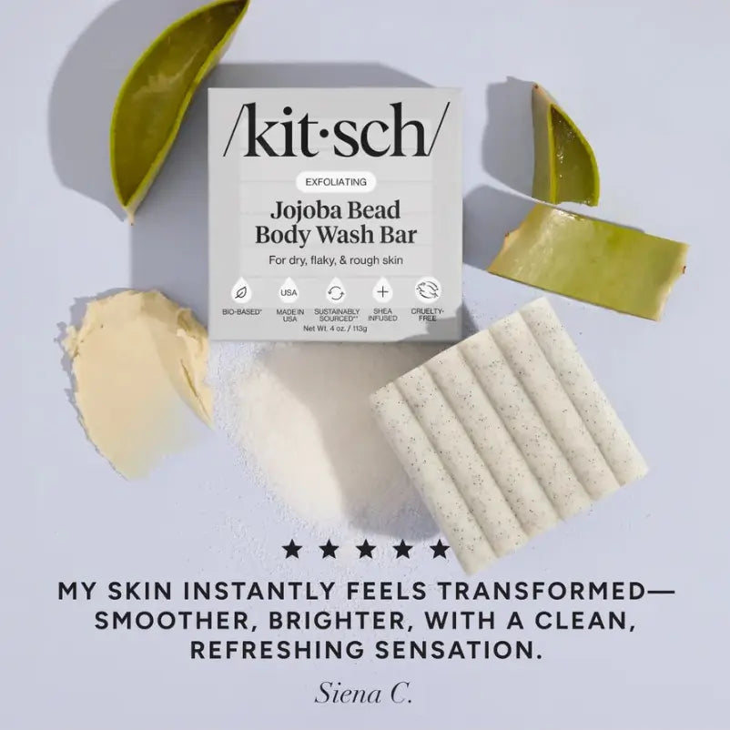 Kit Sch Exfoliating Jojoba Bead Body Wash Bar shown with its white packaging box, surrounded by sliced jojoba plant material and several cylindrical body wash bars. Text at the bottom reads, 'MY SKIN INSTANTLY FEELS TRANSFORMED—SMOOTHER, BRIGHTER, WITH A CLEAN, REFRESHING SENSATION.