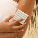 Person using a white exfoliating pad on their arm with a blurred natural background