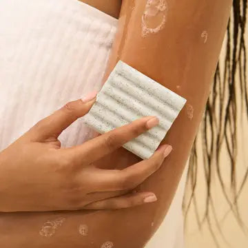 Person using a white exfoliating pad on their arm with a blurred natural background