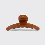 A single large hair claw clip in a glossy amber-brown color, photographed from the side against a light gray background.