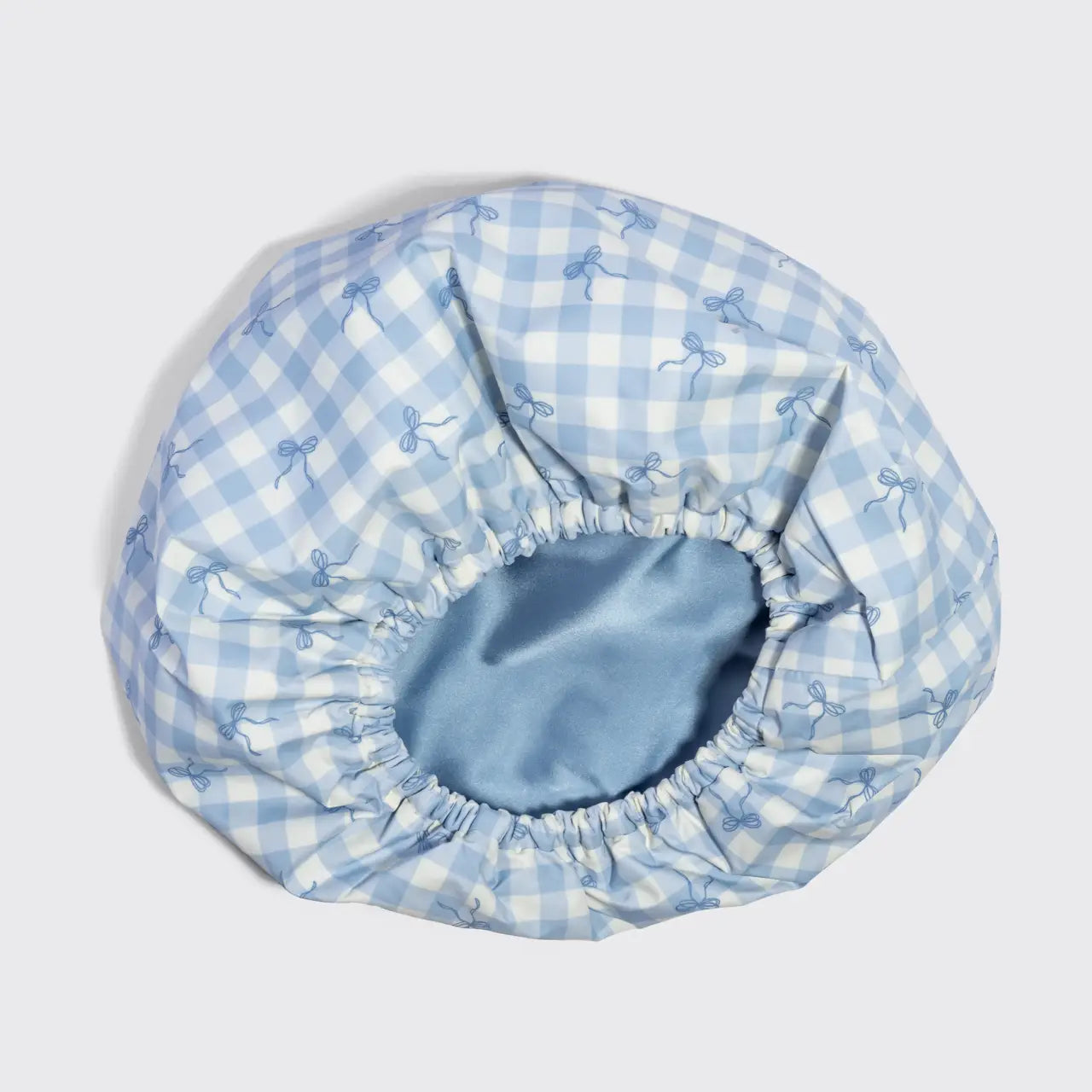 A light blue and white gingham bonnet with a subtle bow pattern, featuring an elasticized blue satin inner band, shown on a neutral light gray background.