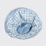A light blue and white gingham bonnet with a subtle bow pattern, featuring an elasticized blue satin inner band, shown on a neutral light gray background.