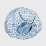 A light blue and white gingham bonnet with a subtle bow pattern, featuring an elasticized blue satin inner band, shown on a neutral light gray background.