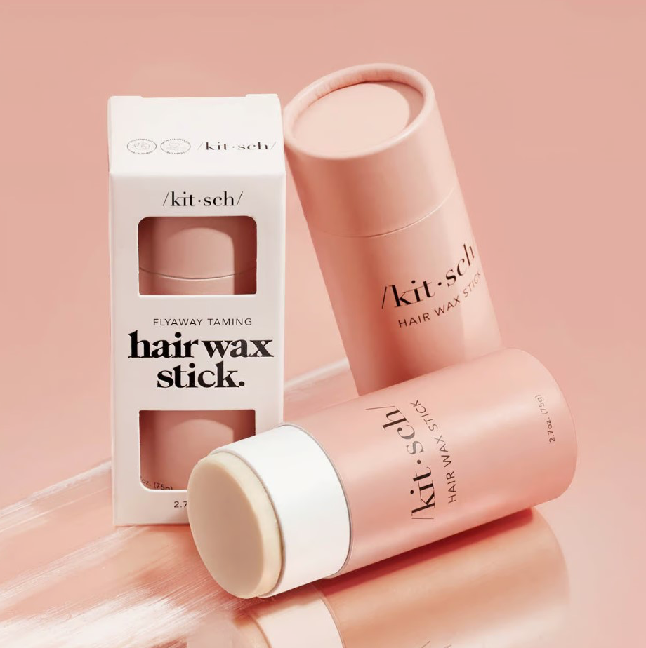 Kit-sch Flyaway Taming Hairwax Stick shown with its white retail box and two pink cylindrical stick containers on a soft pink studio background.