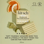 Kit Sch Glycolic Acid Face & Body Wash Bar shown with its beige box, a folded cream-colored soap bar, gold and green soap wrappers, orange slices, and a mint wedge on a pale green background. Text at the bottom reads, 'GIFT YOURSELF AMAZING SKIN! THIS BAR IS LITERALLY THE BEST THING YOU CAN DO FOR YOUR SKIN!' with the name Alison below.