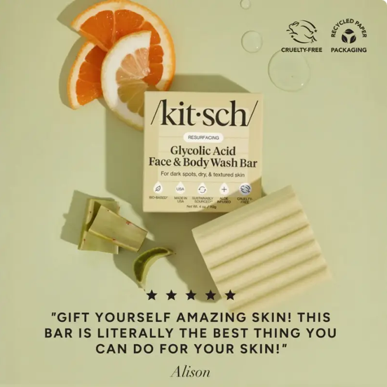 Kit Sch Glycolic Acid Face & Body Wash Bar shown with its beige box, a folded cream-colored soap bar, gold and green soap wrappers, orange slices, and a mint wedge on a pale green background. Text at the bottom reads, 'GIFT YOURSELF AMAZING SKIN! THIS BAR IS LITERALLY THE BEST THING YOU CAN DO FOR YOUR SKIN!' with the name Alison below.