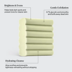 A pale yellow-green rectangular skincare cleanser bar shown on a light gray background with three callout text labels reading Brightens & Evens, Gentle Exfoliation, and Hydrating Cleanse.