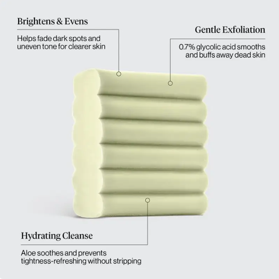 A pale yellow-green rectangular skincare cleanser bar shown on a light gray background with three callout text labels reading Brightens & Evens, Gentle Exfoliation, and Hydrating Cleanse.