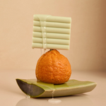 A styled product shot featuring a pale green rectangular block with horizontal ridges, dripping with clear liquid, balanced on top of a textured orange fruit. The fruit sits on a curved green ceramic plate against a warm beige background.