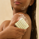 A close-up lifestyle shot of a woman with braided hair applying a pale yellow bar of soap with lather foam to her shoulder, set against a warm beige background.