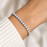 A close-up of a white tennis bracelet with round-cut clear stones set in a silver-toned band, worn on a wrist over a cream ribbed knit sweater.