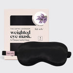 A black satin weighted eye mask displayed in front of its light pink packaging box, which features lavender imagery and the brand name kit.sch.