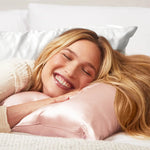 Woman lying on a pink pillow with a smile, surrounded by soft bedding.