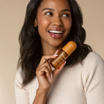 A smiling woman with dark hair holds a Kitsch Caramel Cream Hair Perfume against a warm beige background.
