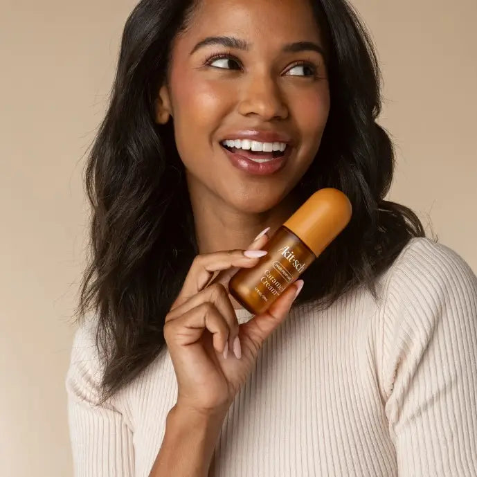 A smiling woman with dark hair holds a Kitsch Caramel Cream Hair Perfume against a warm beige background.