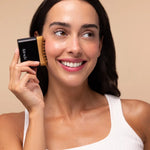 A woman with dark hair and a white tank top holds a black and bamboo facial cleansing brush against her cheek, smiling. The background is a solid beige studio backdrop.