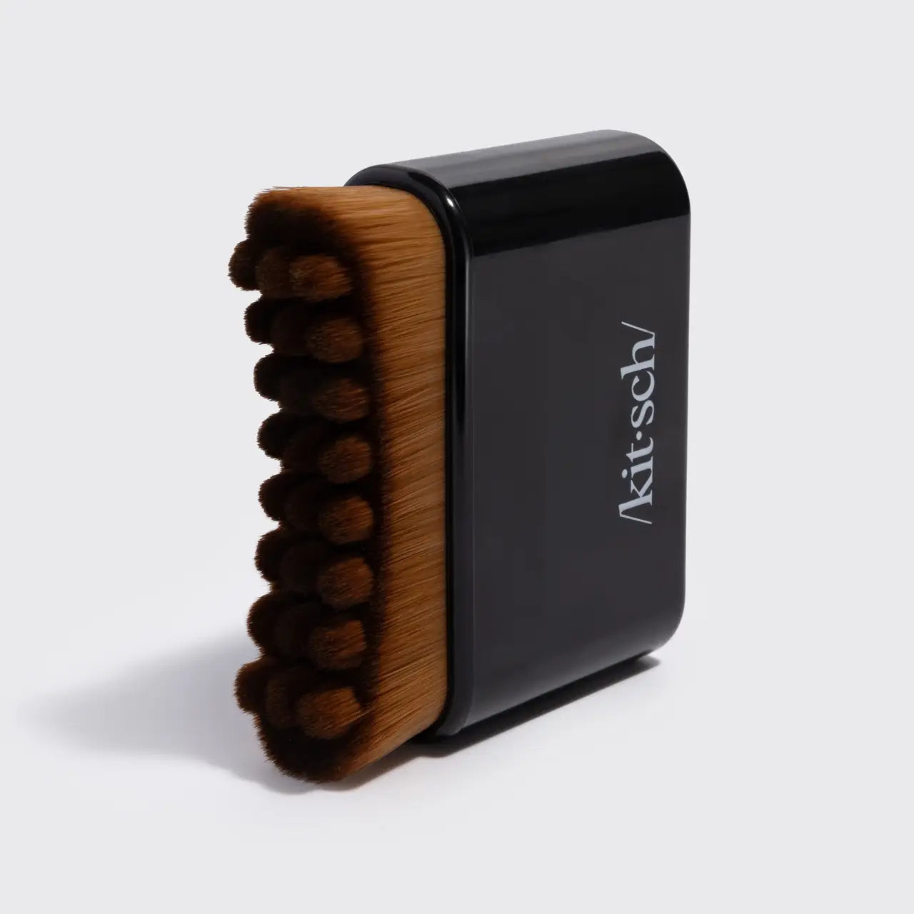 A Kit-sch hand brush with brown bristles and a black handle, standing upright on a light gray background.