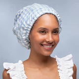 A smiling woman wearing a light blue and white gingham bonnet, photographed against a soft light gray studio background.