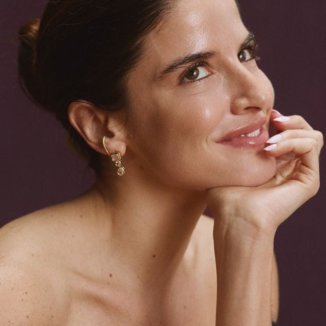 Woman wearing gold three-stone dangle earrings with clear CZ stones in rectangular, round, and pear shapes.