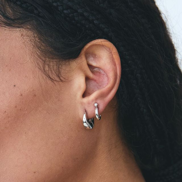 Woman wearing polished silver chunky huggie hoop earrings as part of a layered ear stack.